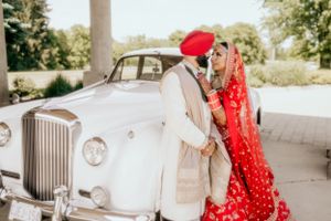 Newlywed couple in traditional Sikh wedding attire pose beside classic white Rolls Royce.