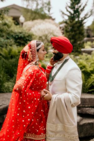 Traditional Sikh wedding couple in red and white outfits pose in a lush garden setting.