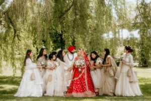 Wedding party in cream dresses gathers around couple in traditional Sikh attire under willow trees.