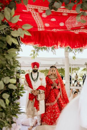 Sikh wedding couple in traditional red attire standing under a decorated mandap with greenery.
