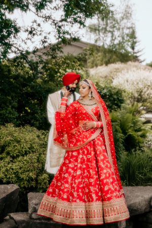 Sikh wedding couple poses in traditional red and white attire among spring blossoms.