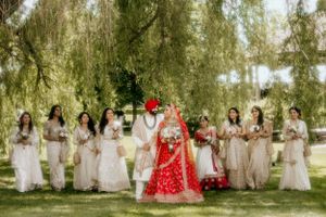 Wedding party poses under drooping willow trees with bride in vibrant red traditional Indian lehenga surrounded by bridesmaids.