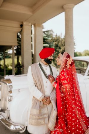 Elegant couple in traditional Sikh wedding attire standing by vintage white car under columned portico.