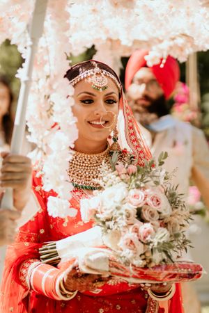 Traditional Indian bride in red lehenga holding bouquet under floral canopy at outdoor ceremony.