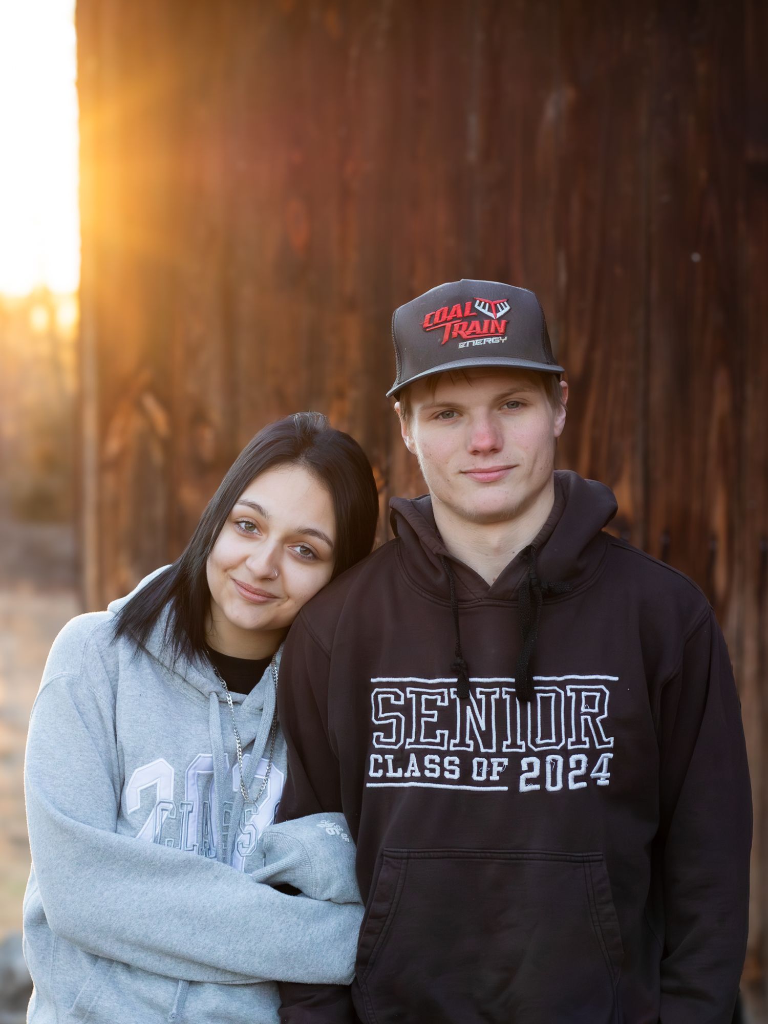 Husband and wife smiling at the camera hugging in front of the lake a fall trees.