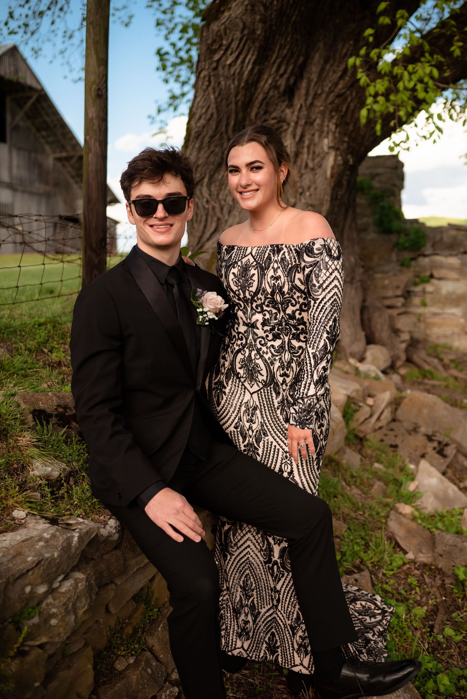 Couple posing together on stone wall wearing formal black and patterned attire in outdoor setting.