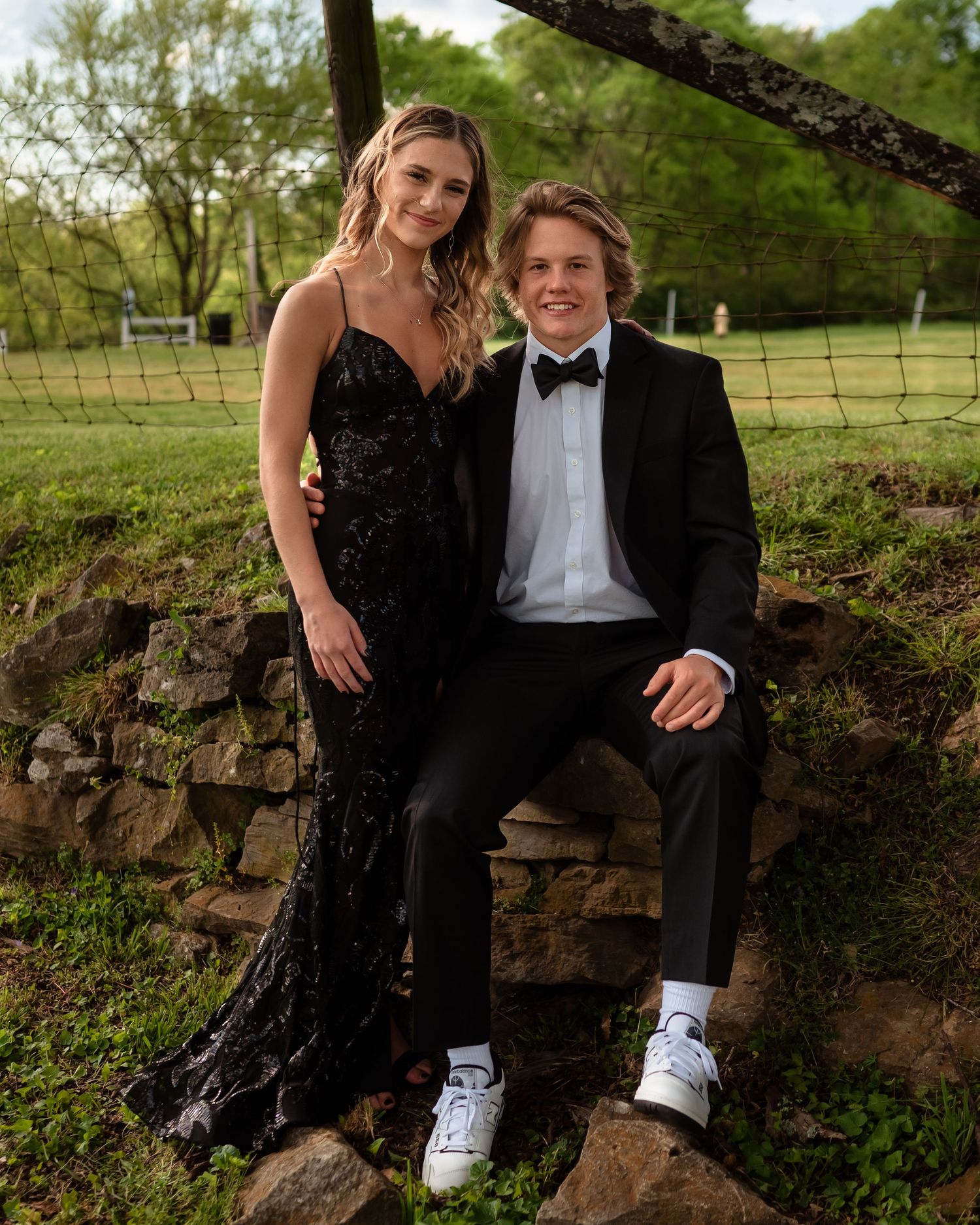 Couple in formal attire sitting on stone wall outdoors with unique sneaker and dress shoe combination.