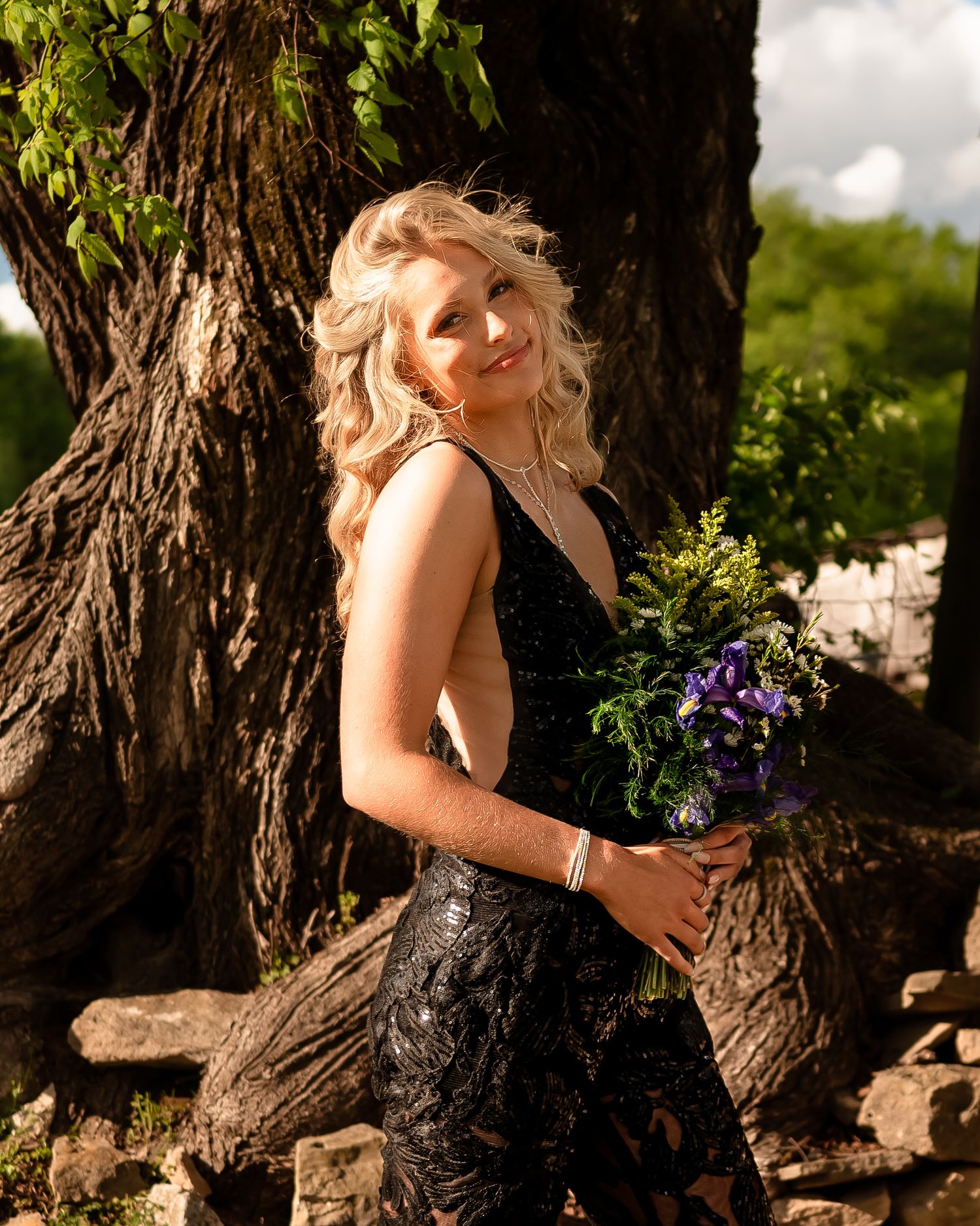 Person in black dress posing artistically against tree trunk in outdoor garden setting.