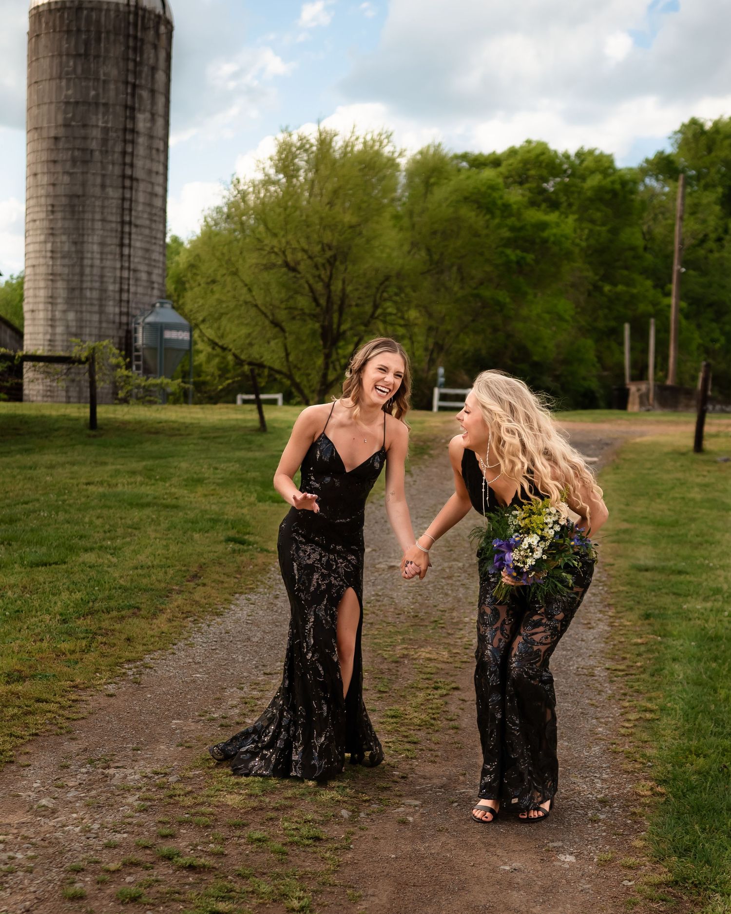 Two people in black formal dresses posing playfully near old farm silos.