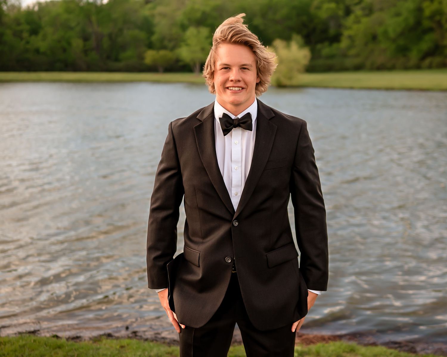 Person in formal black tuxedo poses by lake during outdoor photo session.