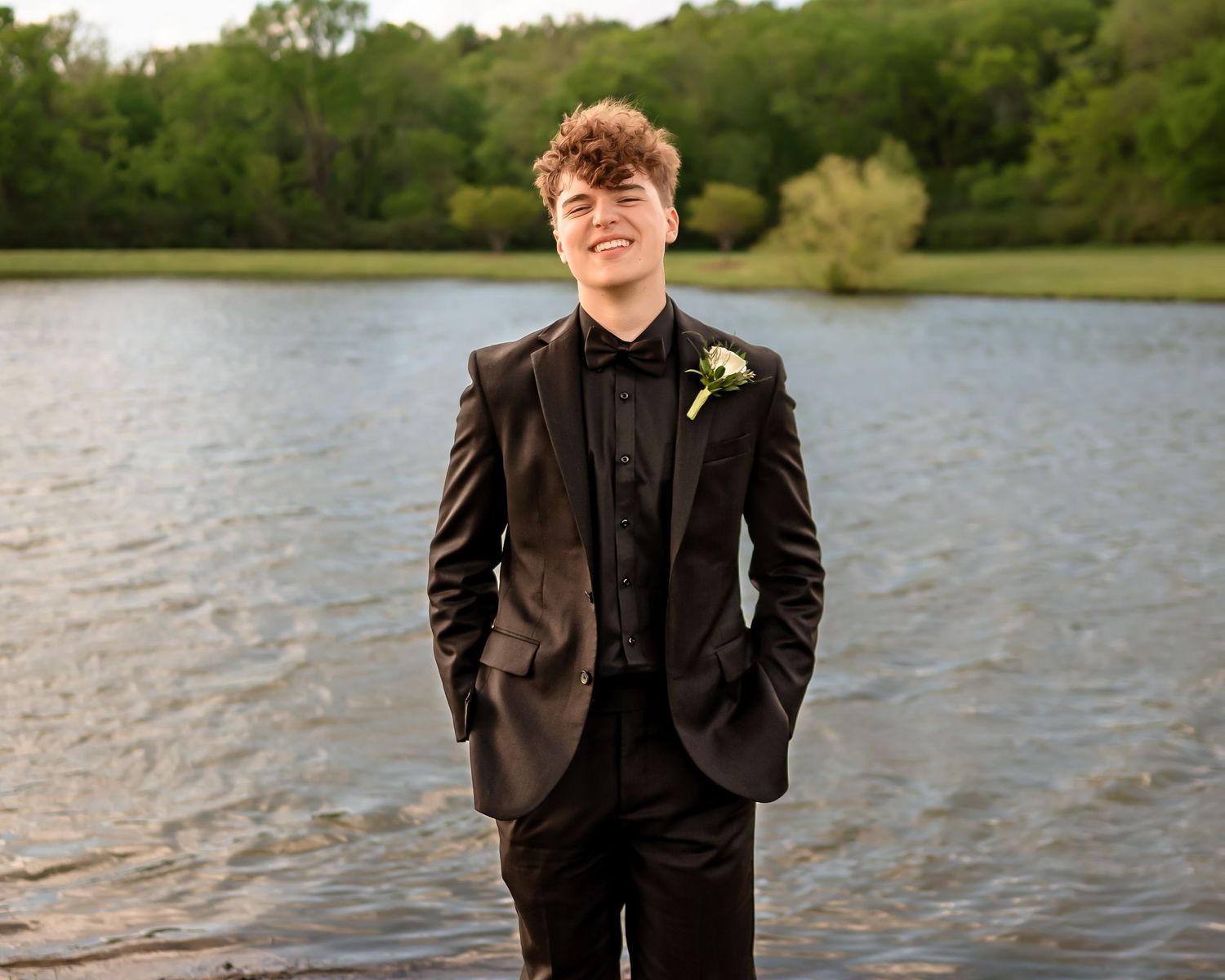 Person in classic black tuxedo smiles during lakeside formal photo session.