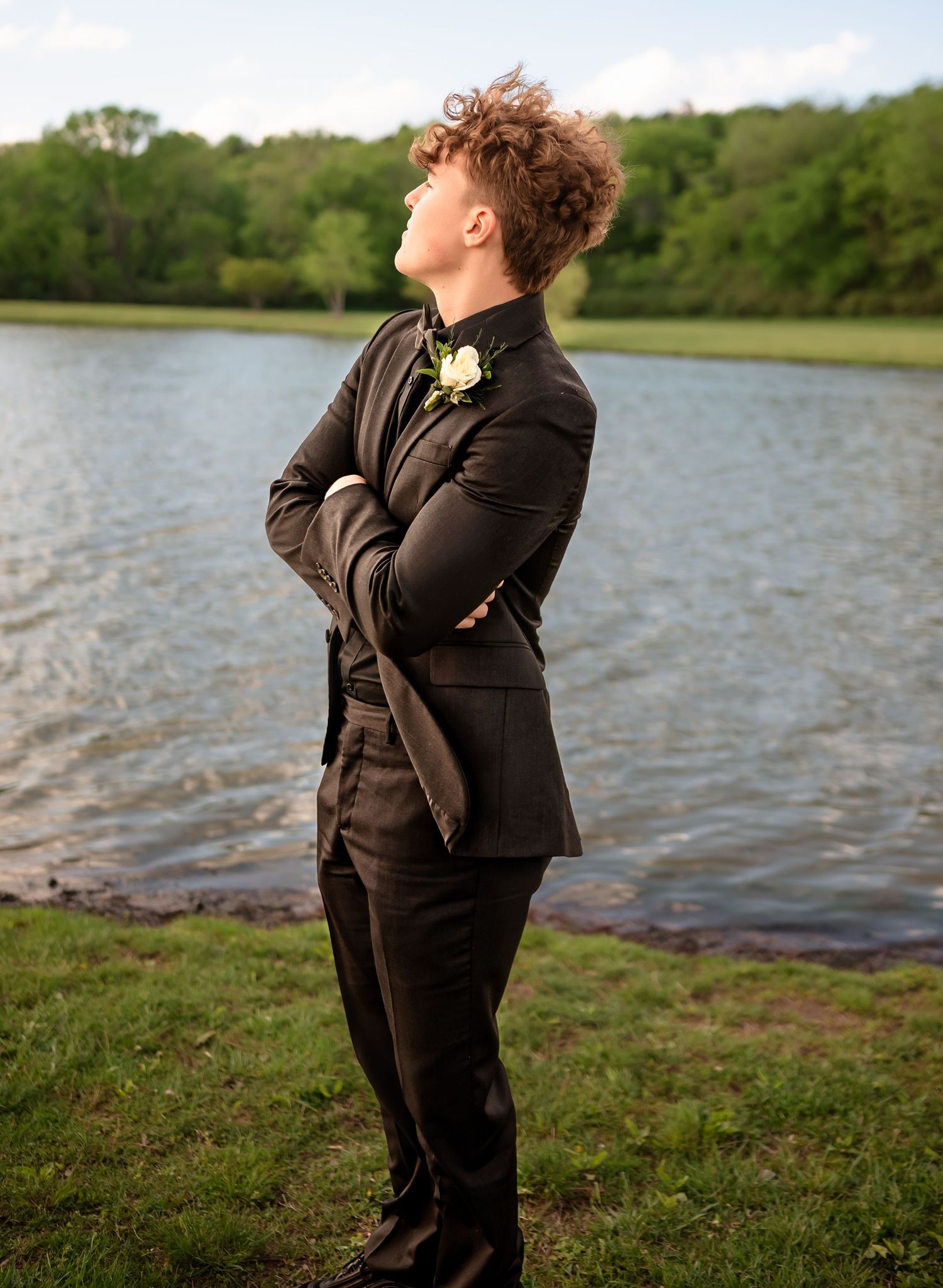 Person in black tuxedo strikes confident pose by lake during formal photoshoot.