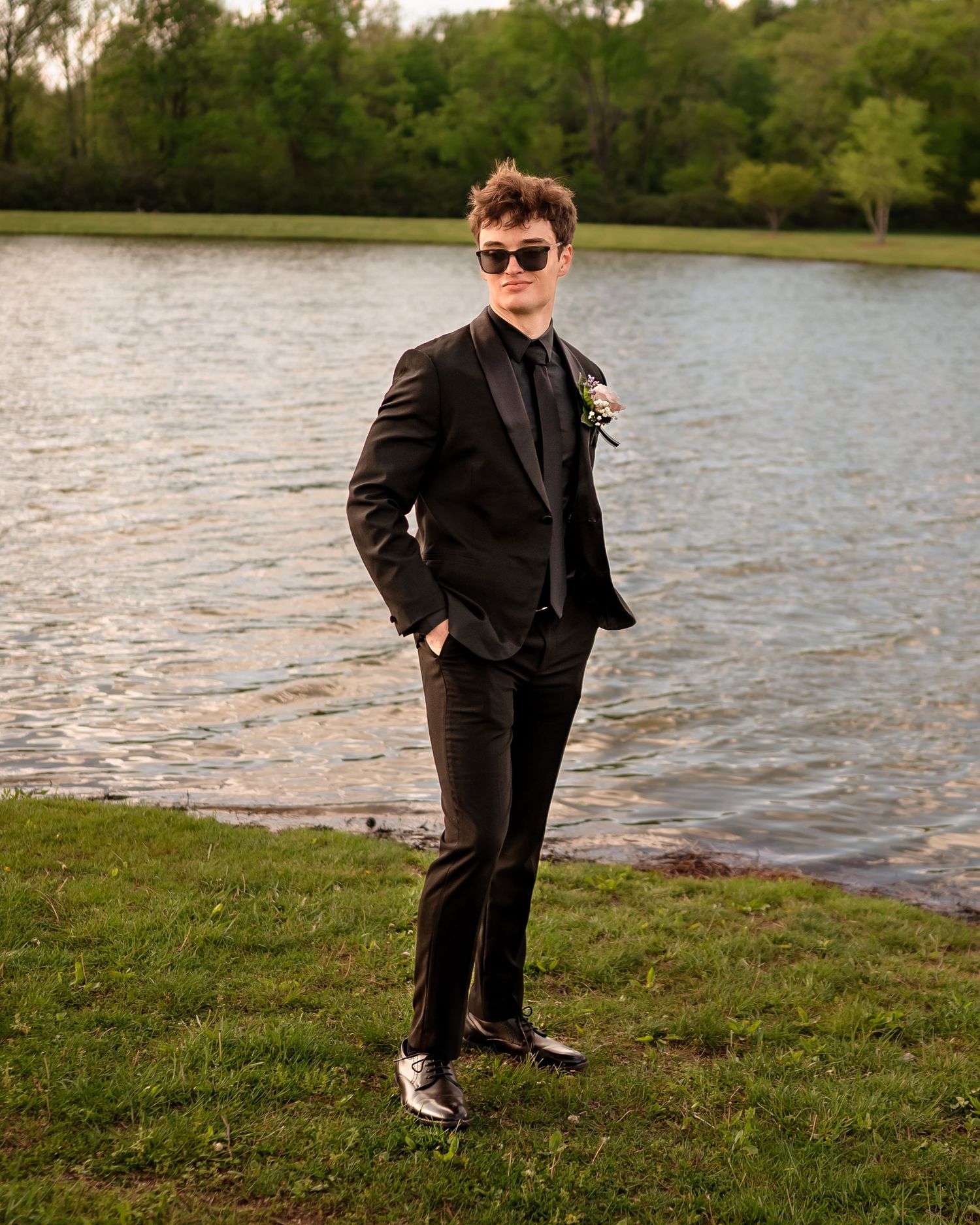 A person in formal black attire poses playfully by a lake during a photo session.