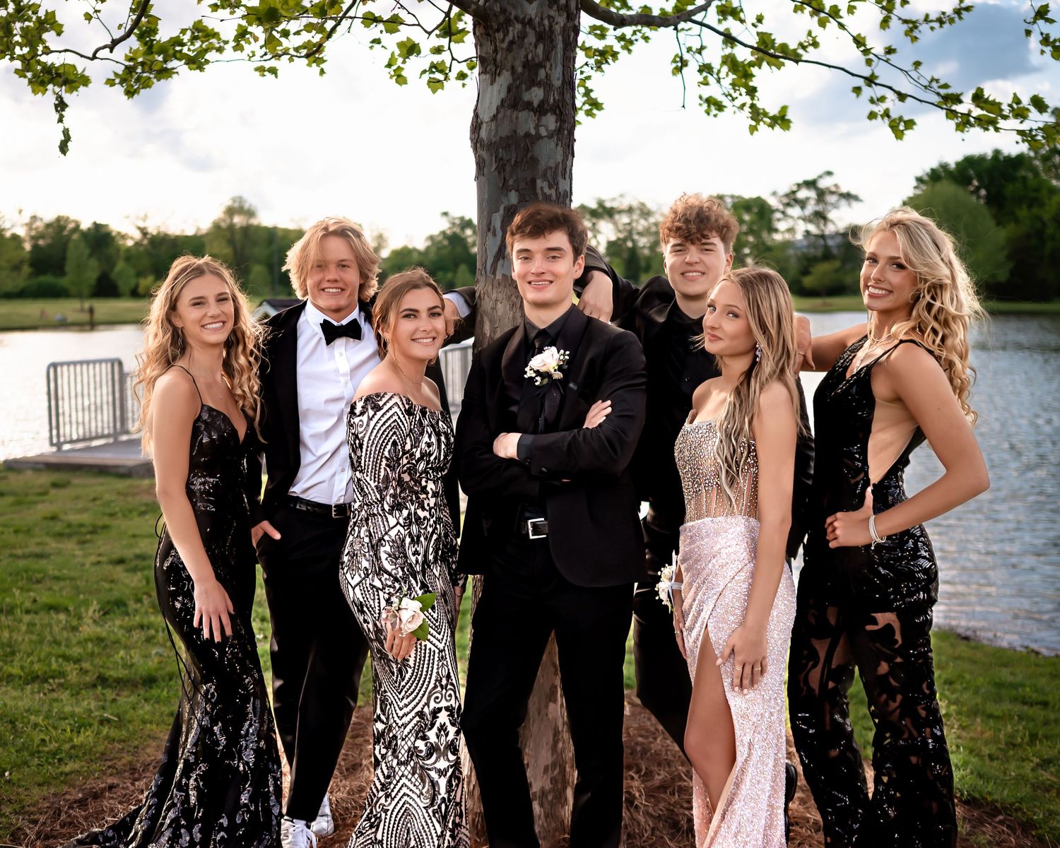 A group in formal dresses and suits poses together by a lakeside for an event photo.