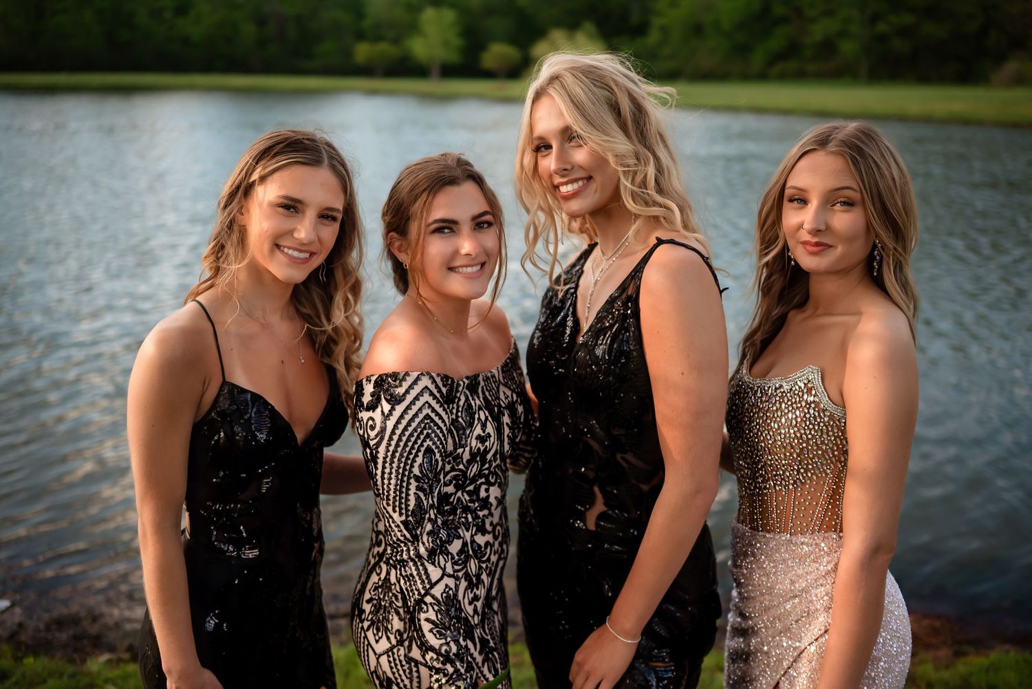 Four friends in elegant dresses pose by lakeside during sunset.