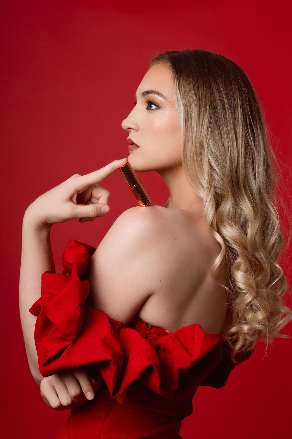 Side-profile of blonde woman in red dress holding lipstick against bold red background – NYC beauty portrait.