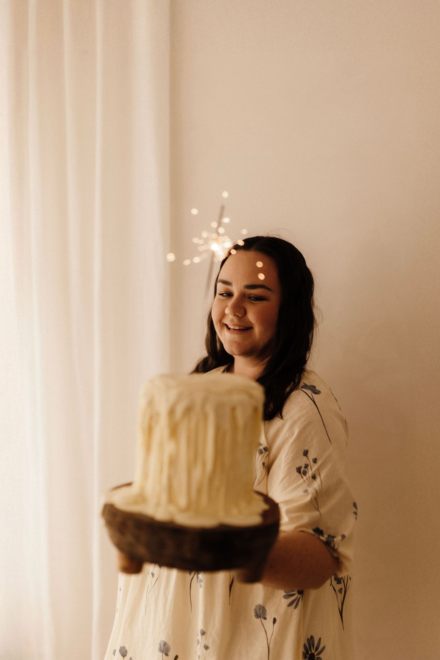 Person in floral dress holding decorated cake with sparklers in soft lighting.