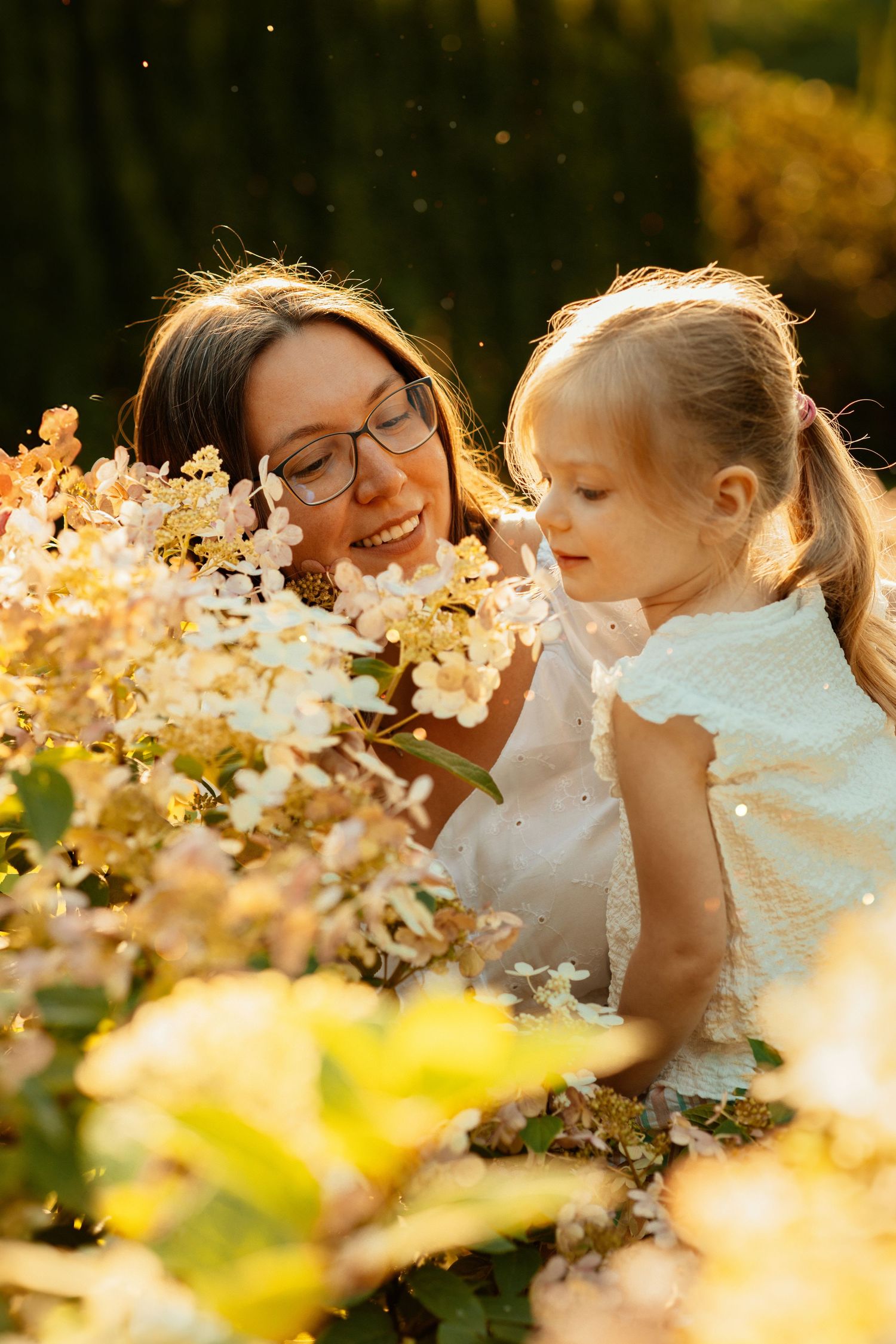a woman in a white dress with a child stands near a flower bush bathed in the rays of the sun