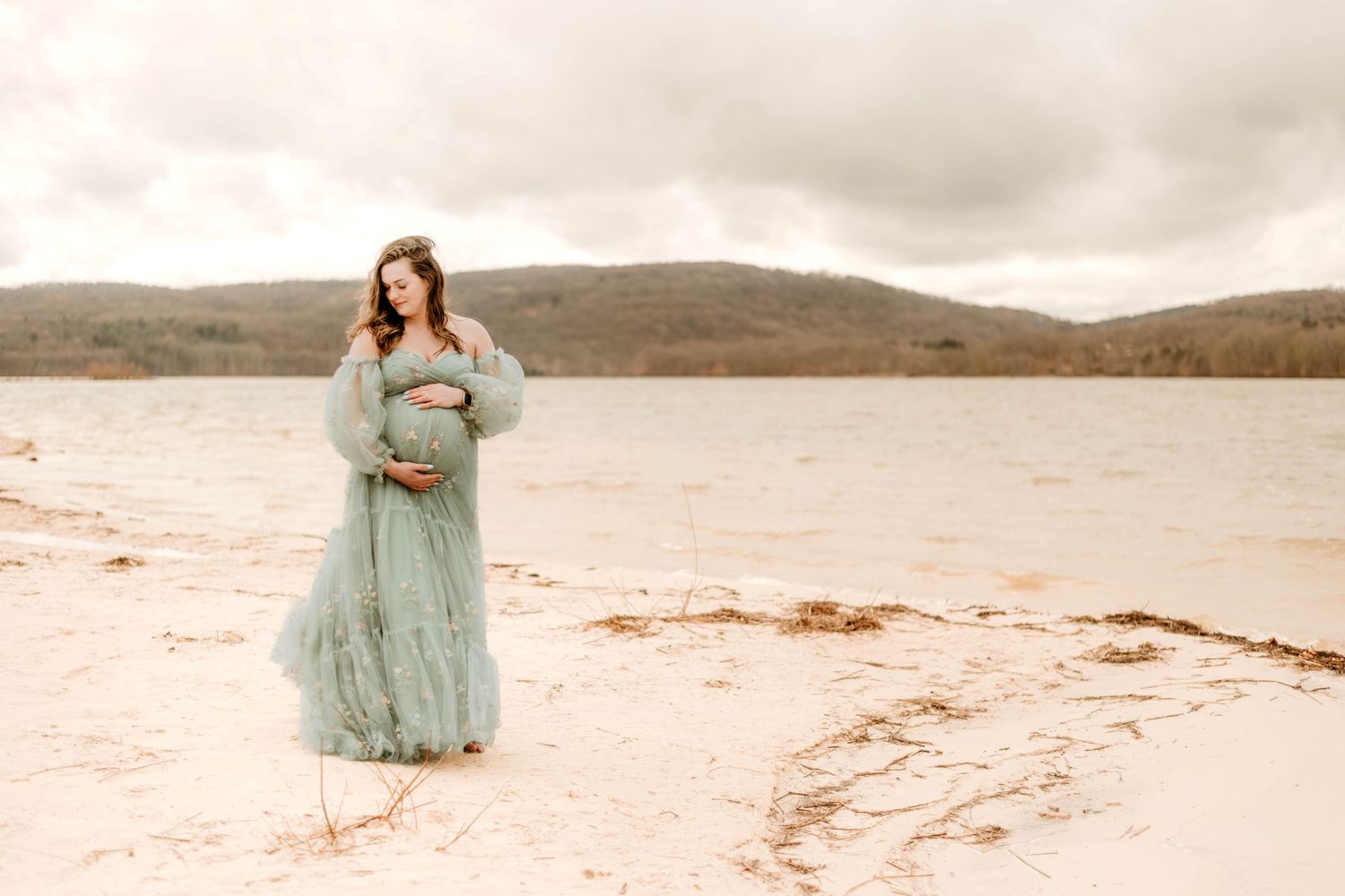 Pregnant woman in teal dress standing on beach shore with mountains in background.