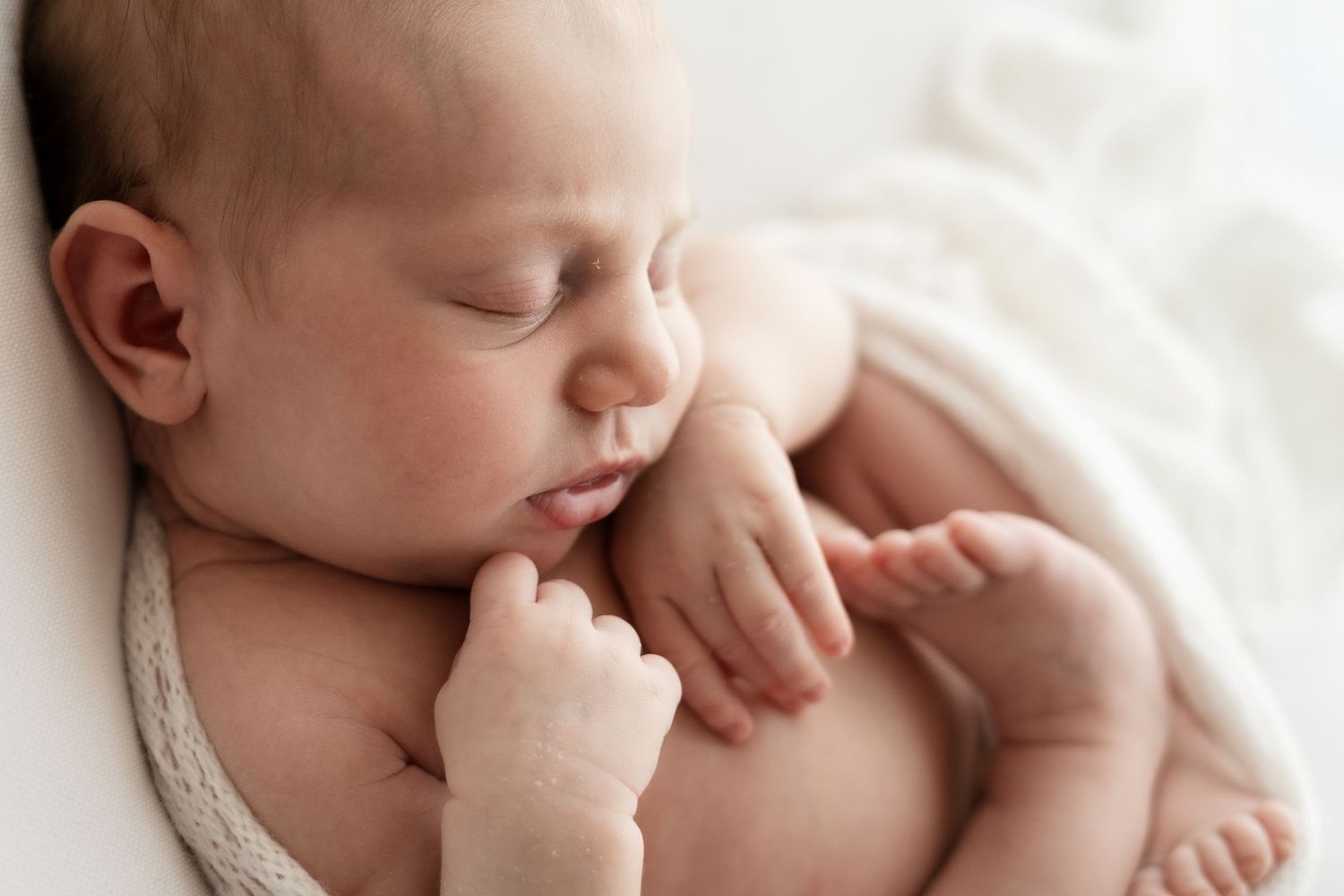Peaceful newborn baby sleeping with tiny hands tucked under chin on white blanket.