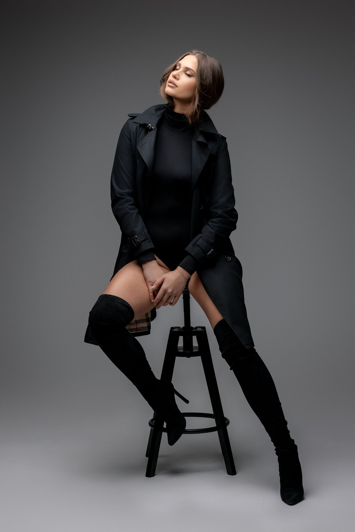 Dramatic fashion portrait with model seated on stool wearing black coat and boots.