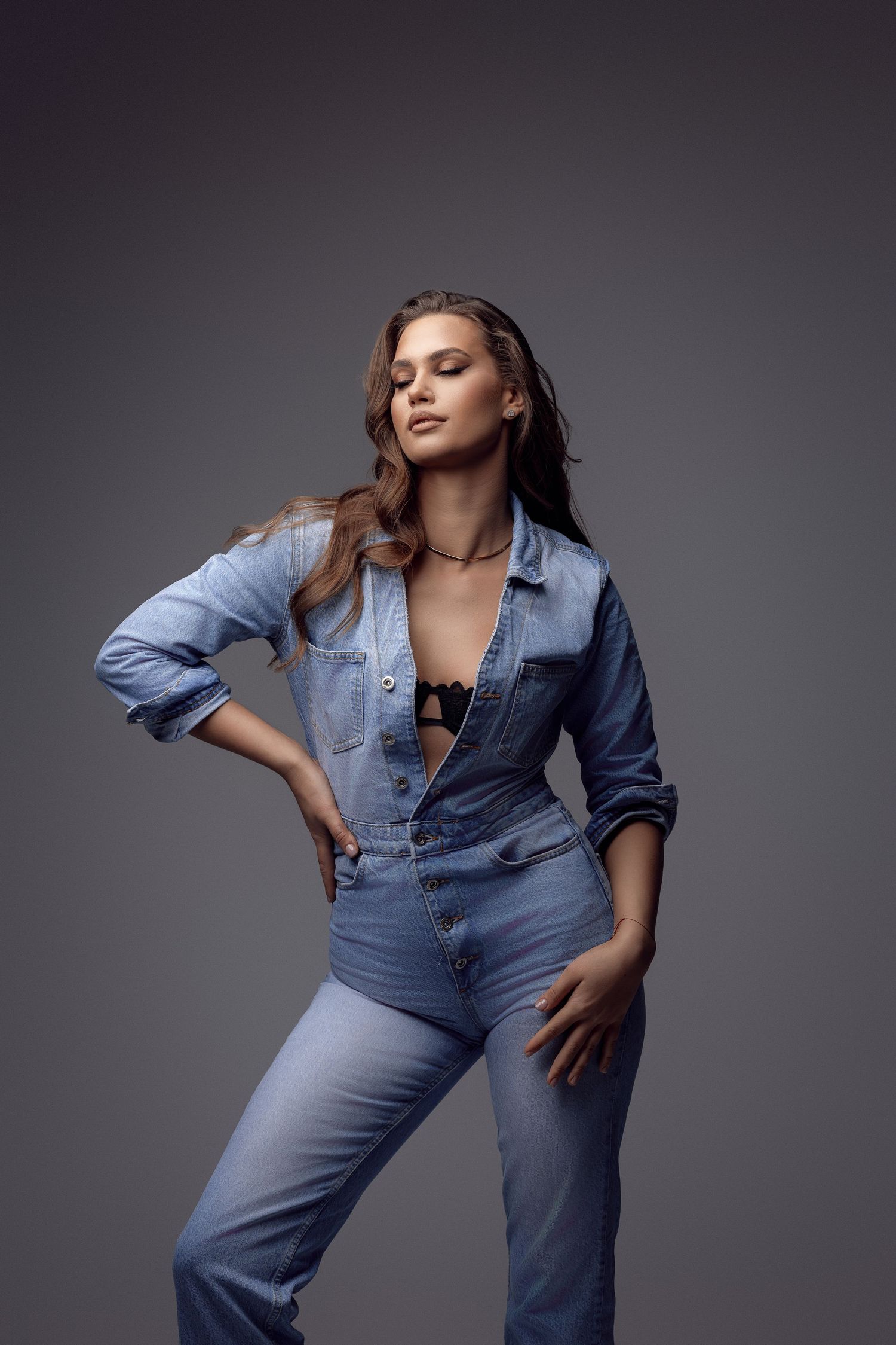 Fashion model poses in casual denim outfit against dark studio background.
