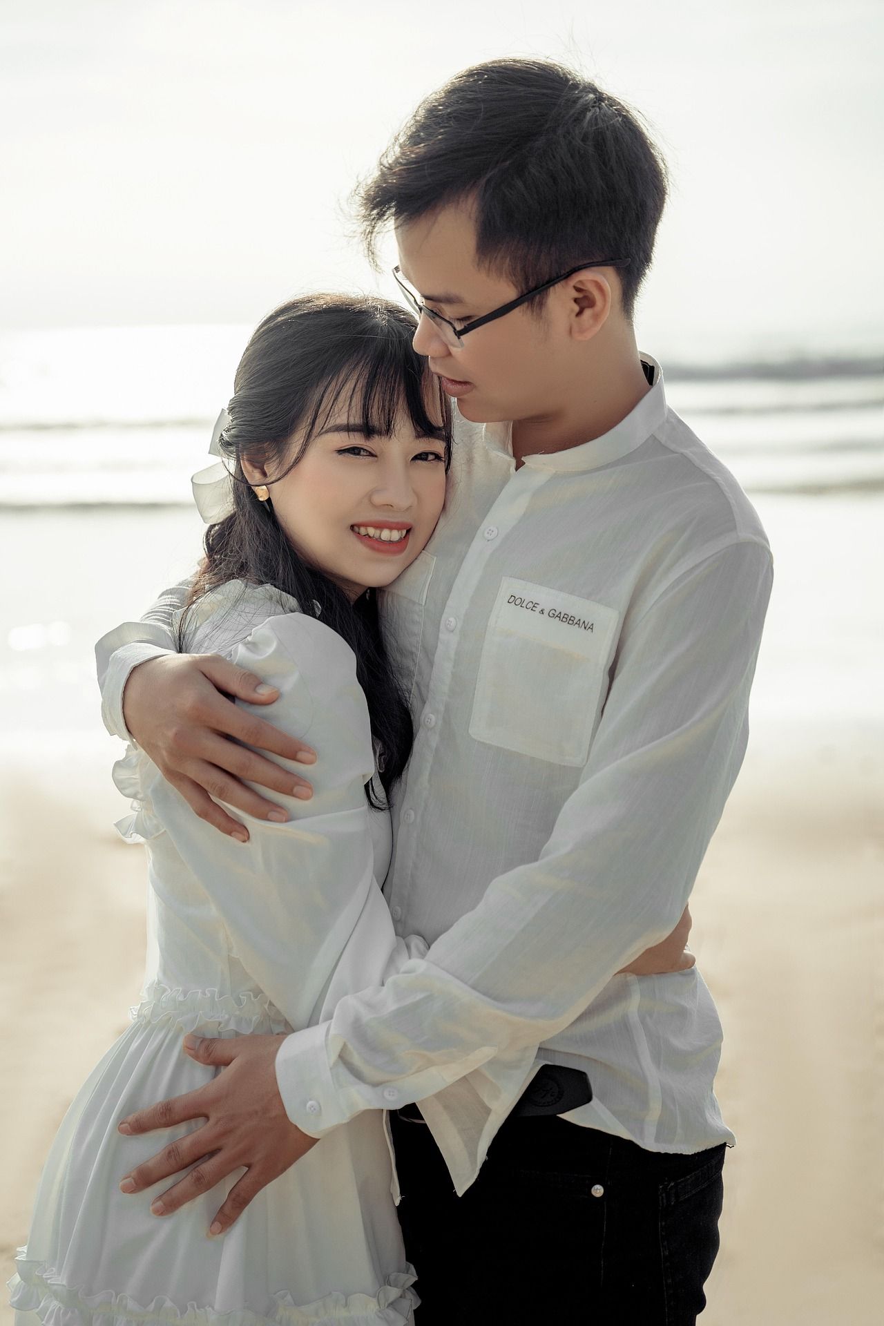 Romantic couple in white outfits embracing on beach during golden hour.