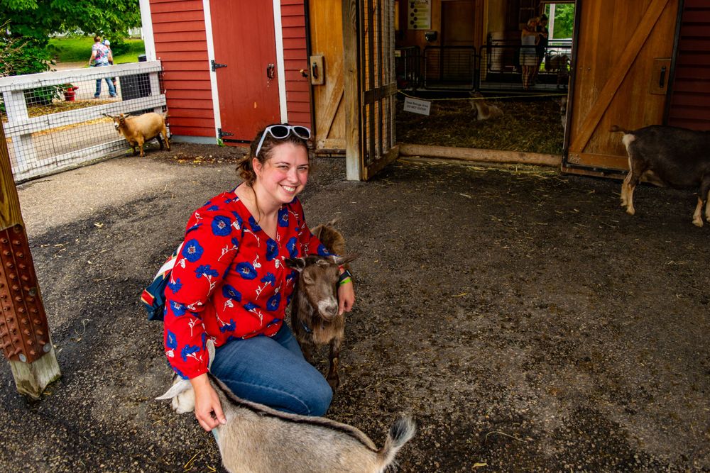 Someone pets a small goat while sitting outside a red barn structure.