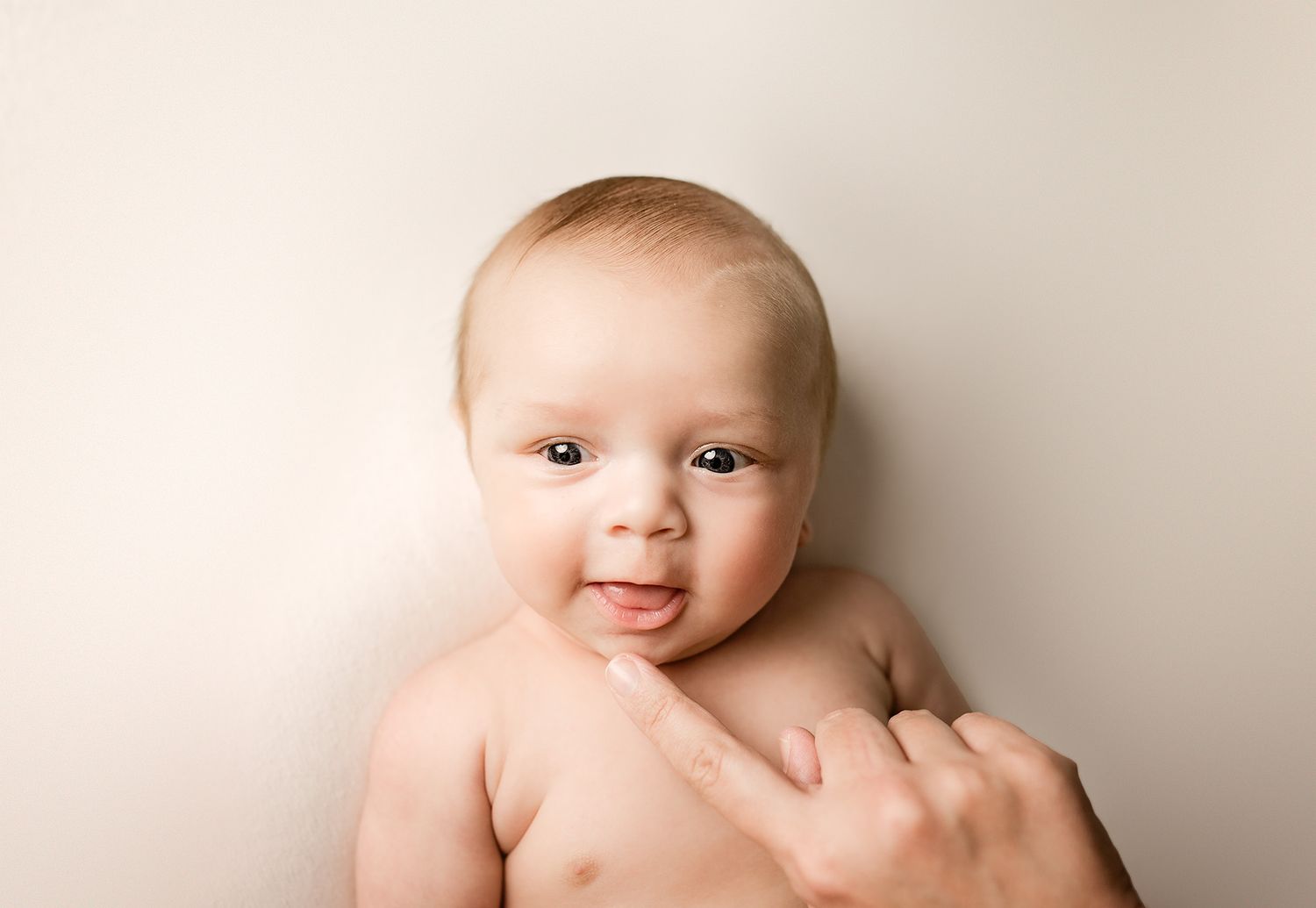 Cute smiling baby held upright against cream background with gentle hand supporting tiny chest.