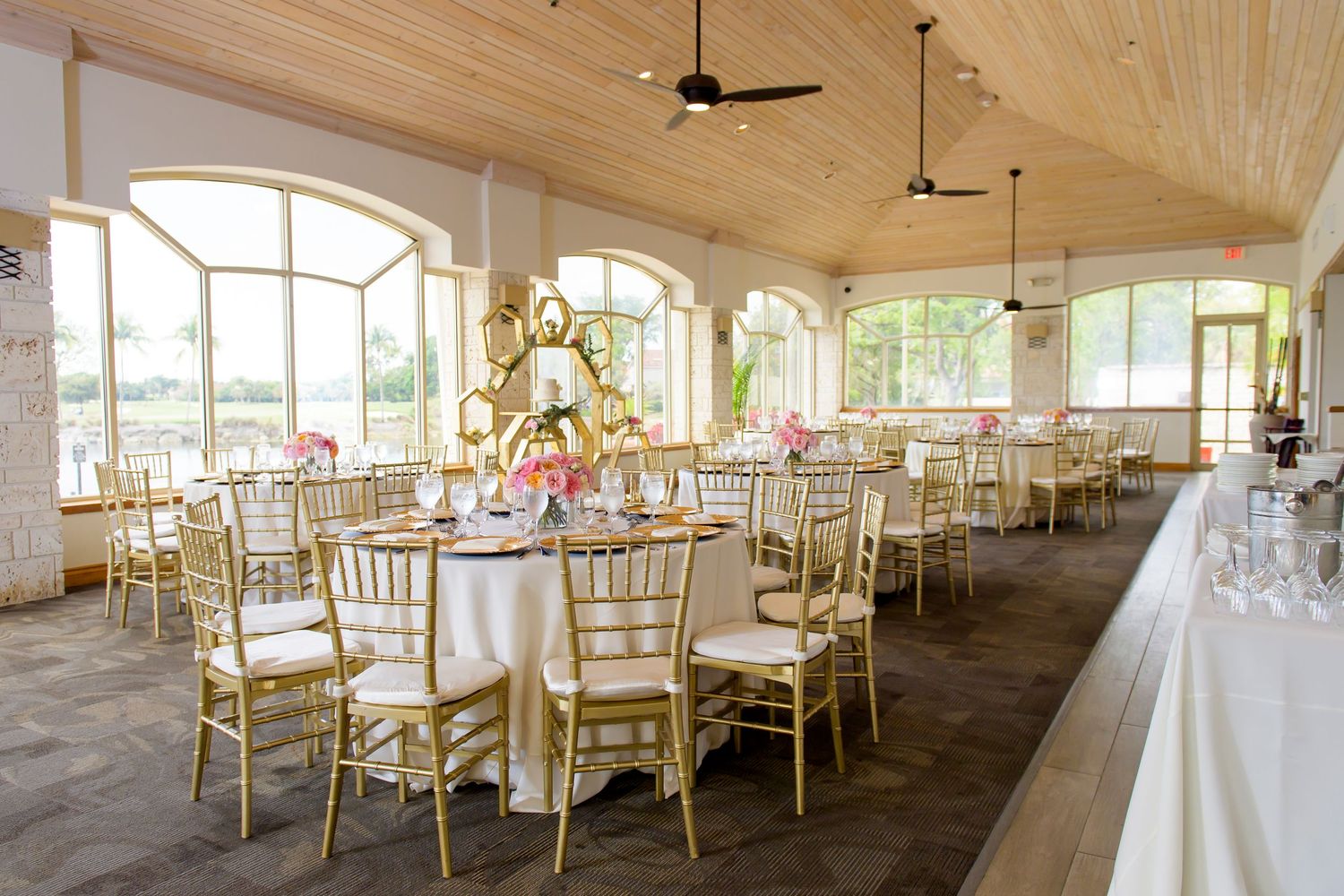 Bright sunlit reception room with gold chairs, white tablecloths, and large arched windows.
