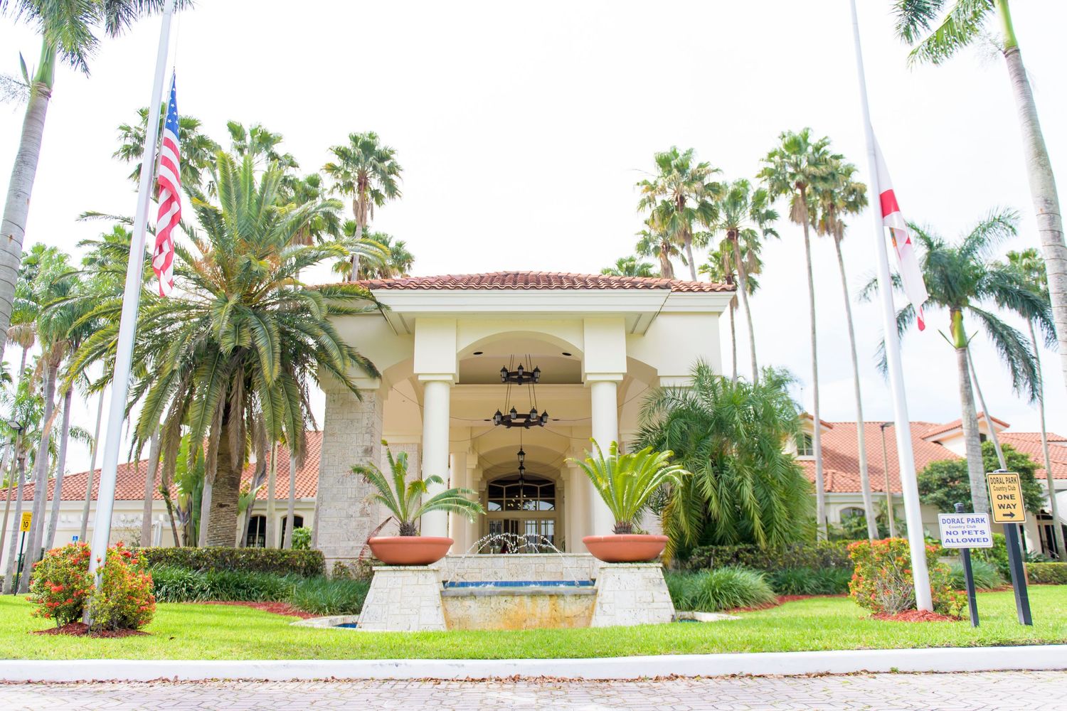 Mediterranean style building entrance with palm trees and American flag in tropical setting.