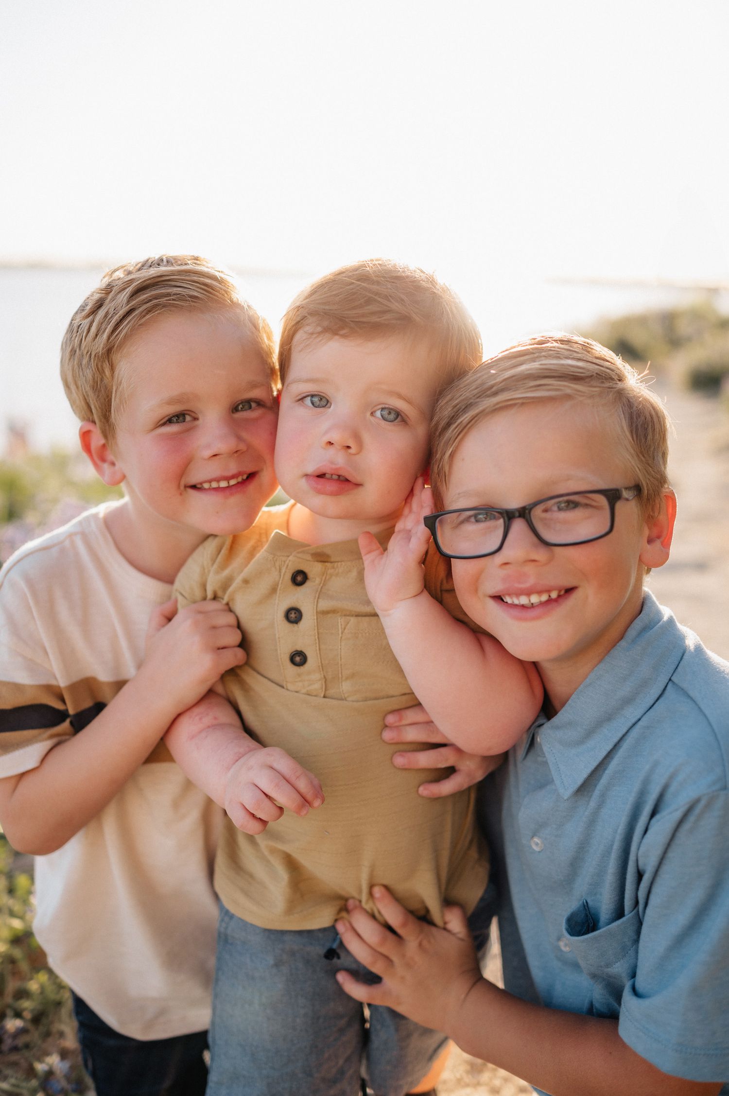 Three siblings share a warm embrace while smiling in golden sunlight.
