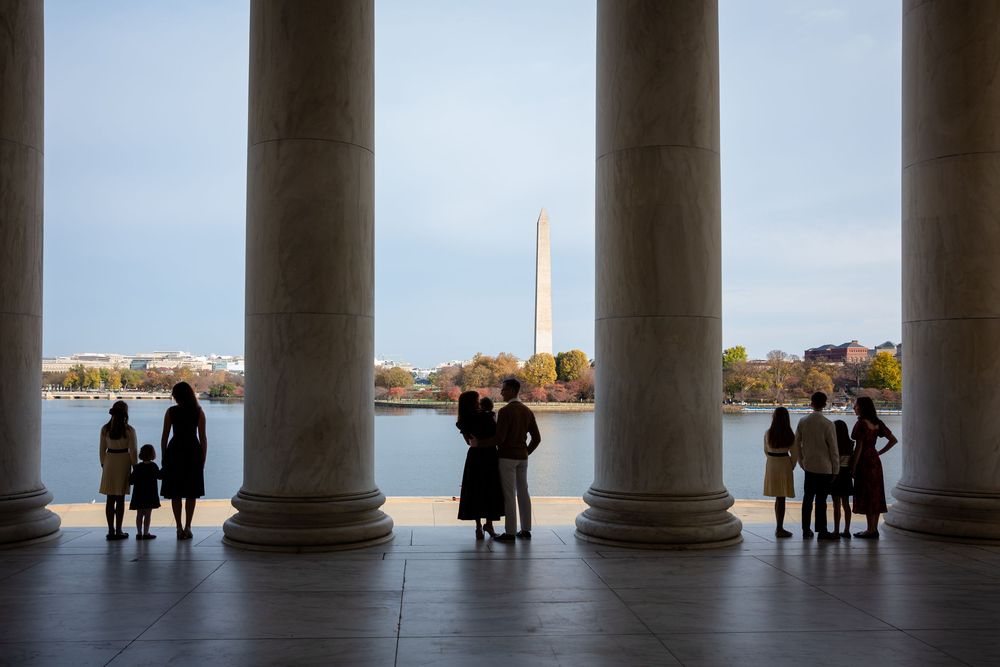 Silhouettes of visitors stand between tall columns at the Jefferson Memorial, with the Washington Monument reflected across the Tidal Basin.