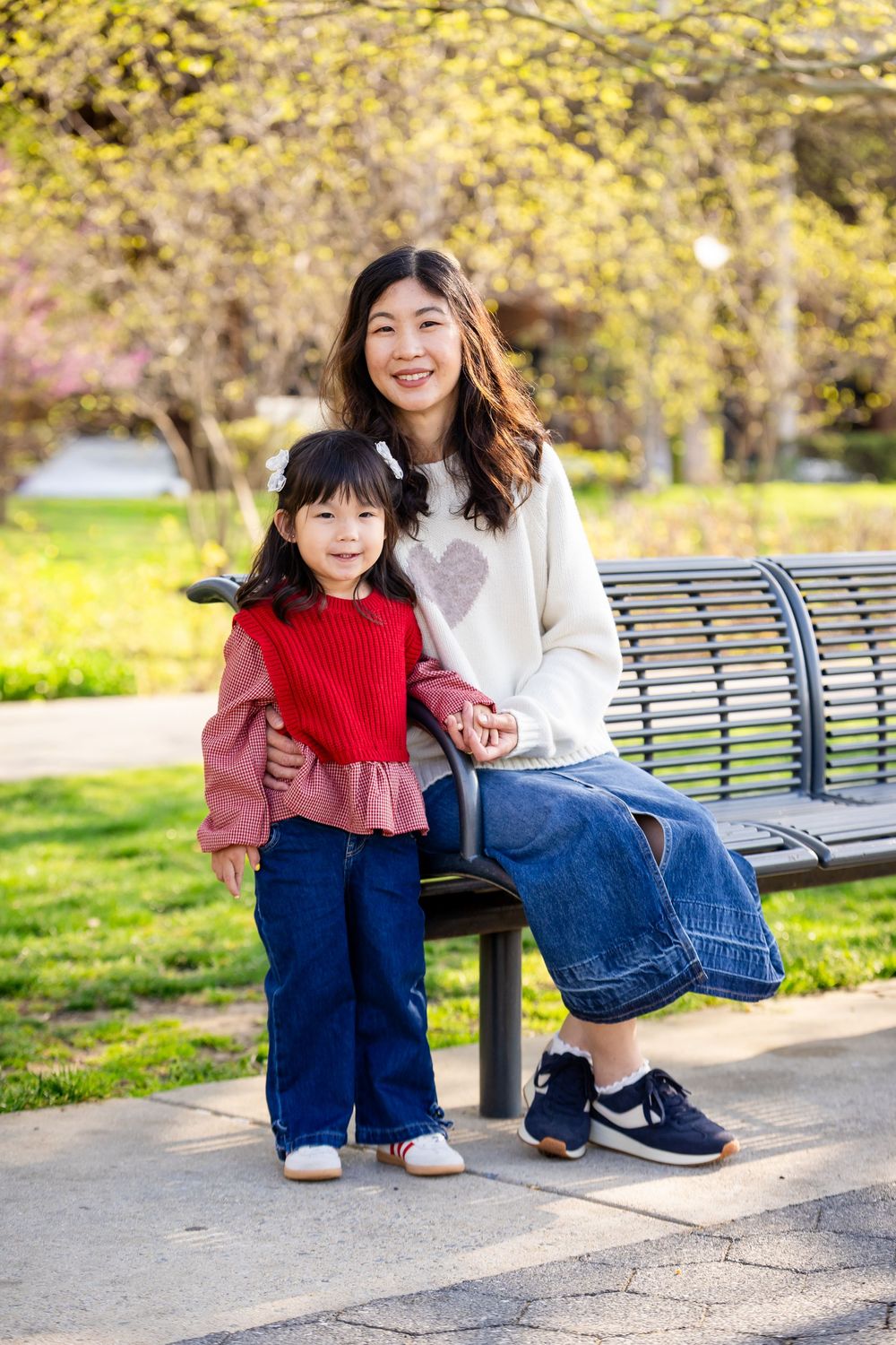 A smiling Asian woman sits on a park bench with a young girl in a red vest, surrounded by spring blooms.