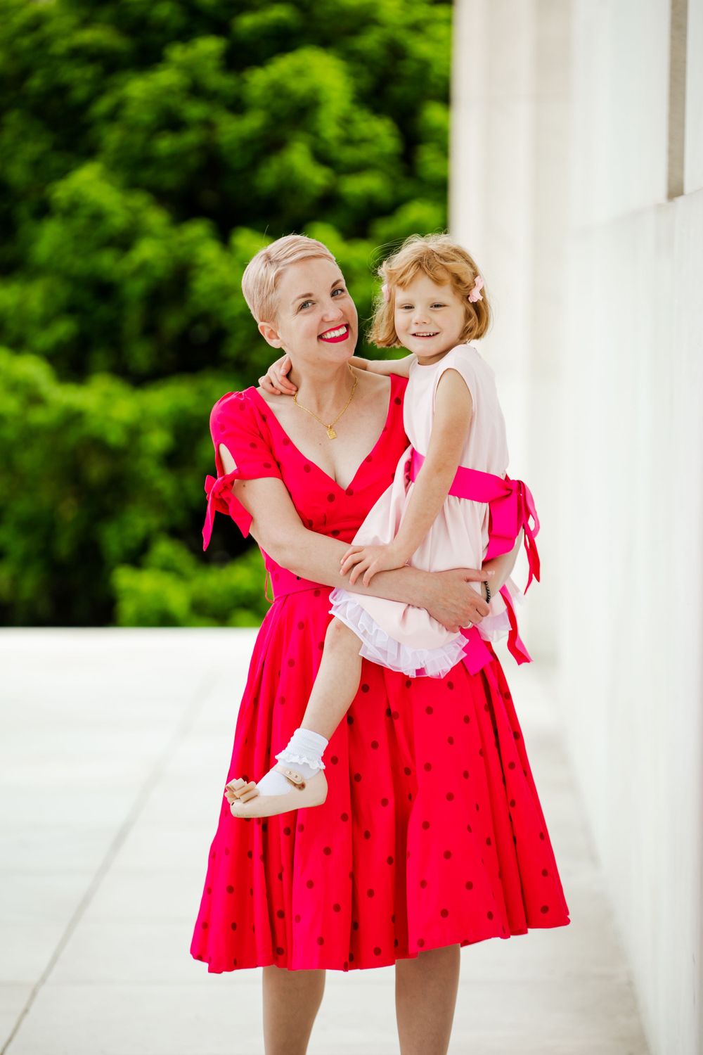 Mom holding daughter both looking at camera with green trees and white marble wall in background, by Justin Baker Photography. Washington DC