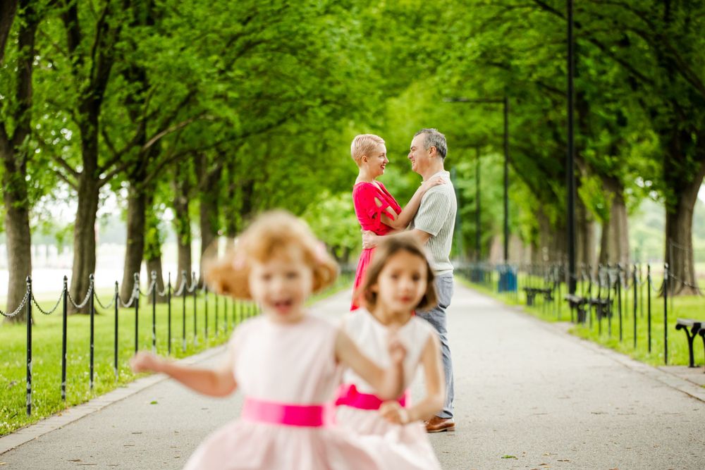 Young children play in foreground as parents dance under tree-lined path in summer park, captured by Justin Baker Photography in DMV.