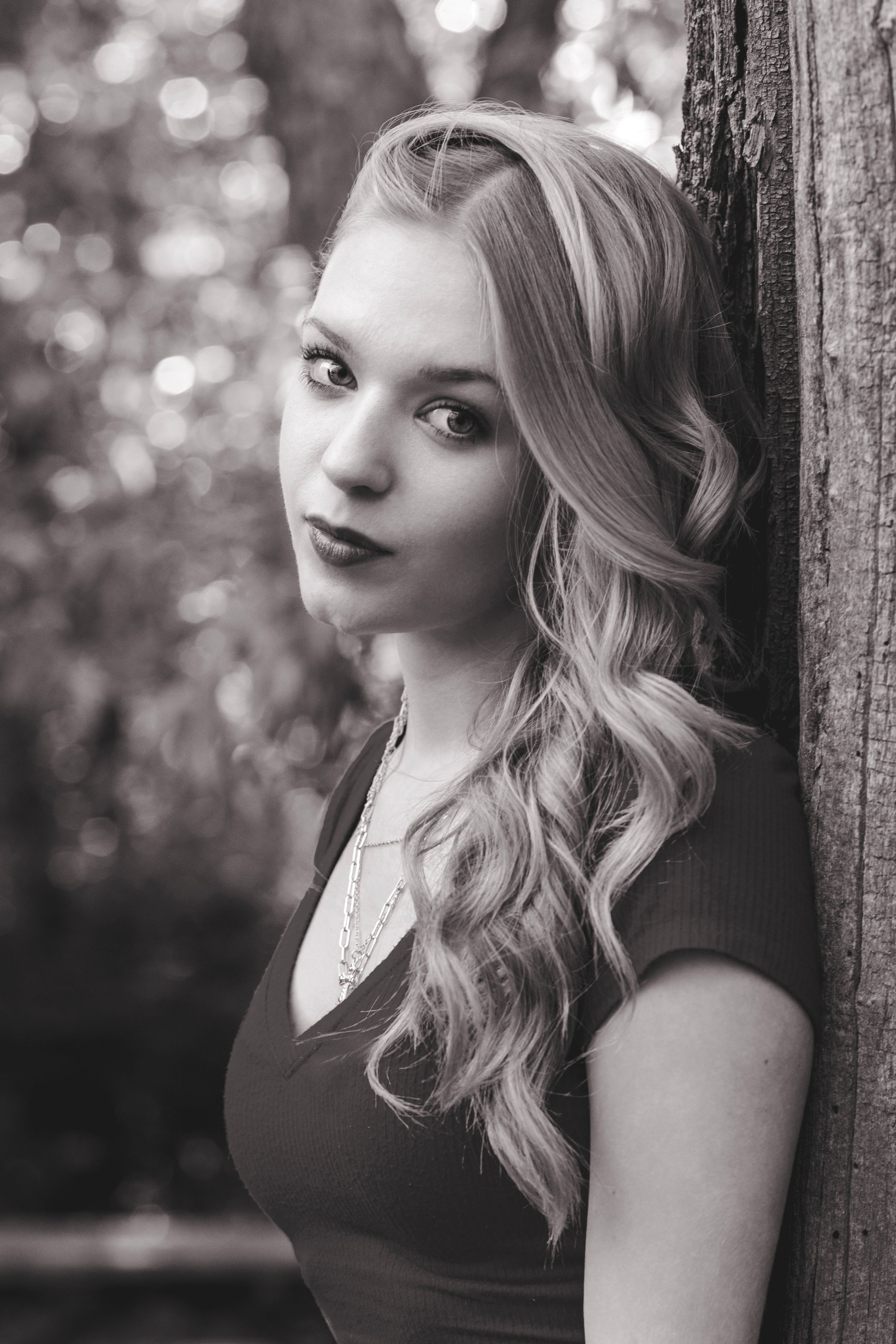 Black and white portrait of a young blonde woman leaning against a wooden post outdoors.