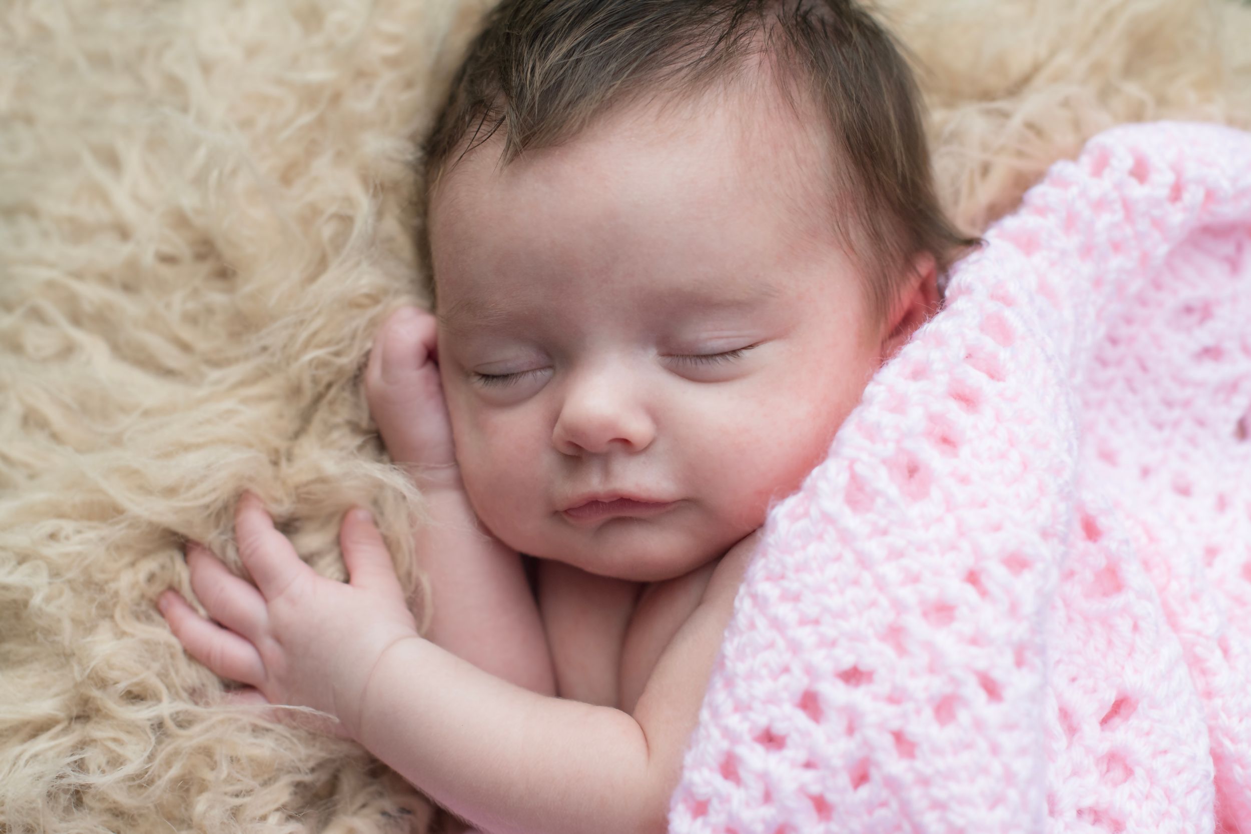 Newborn baby sleeping peacefully on a fluffy cream rug, wrapped in a soft pink crochet blanket.