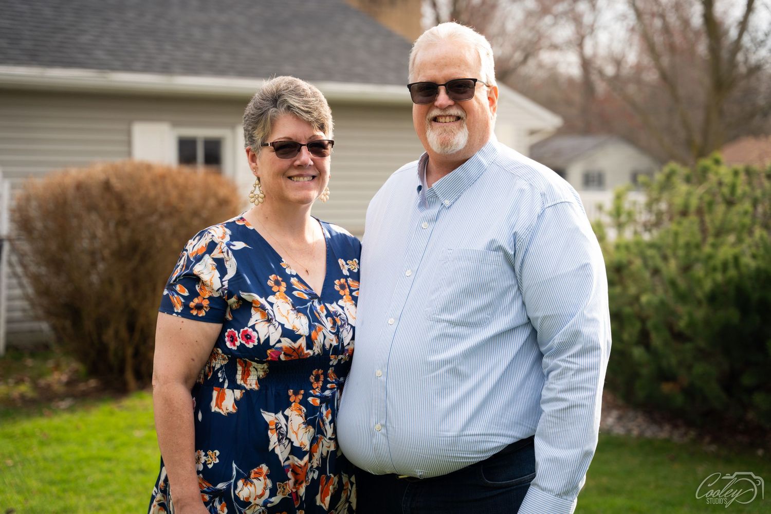 Smiling couple posing outdoors in front of suburban home on overcast day.