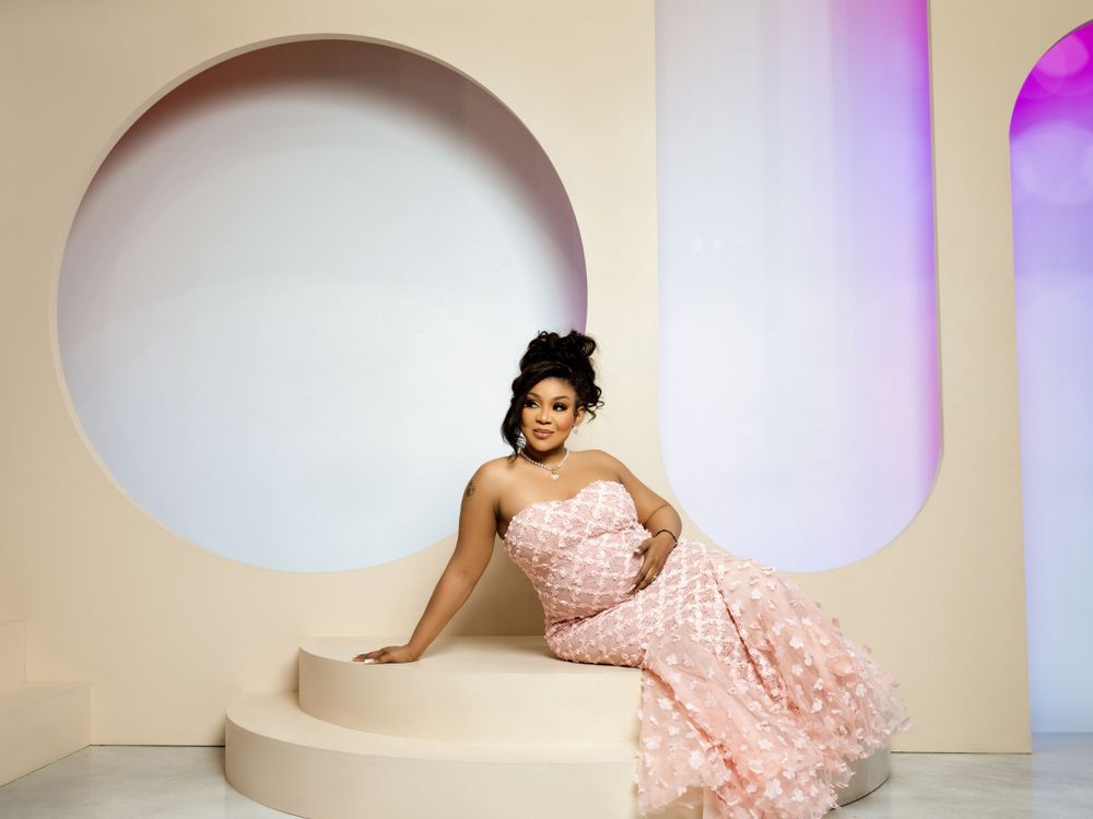A model in a pink strapless ball gown poses elegantly on a curved white platform against a modern architectural backdrop.