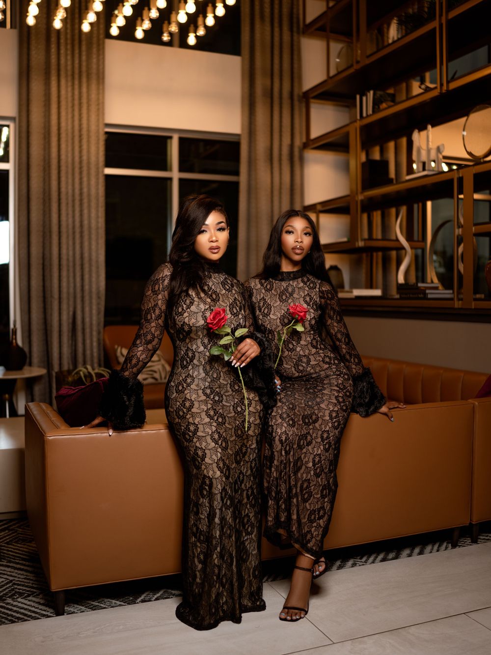 Two women in matching lace dresses pose in an upscale lounge with warm lighting.