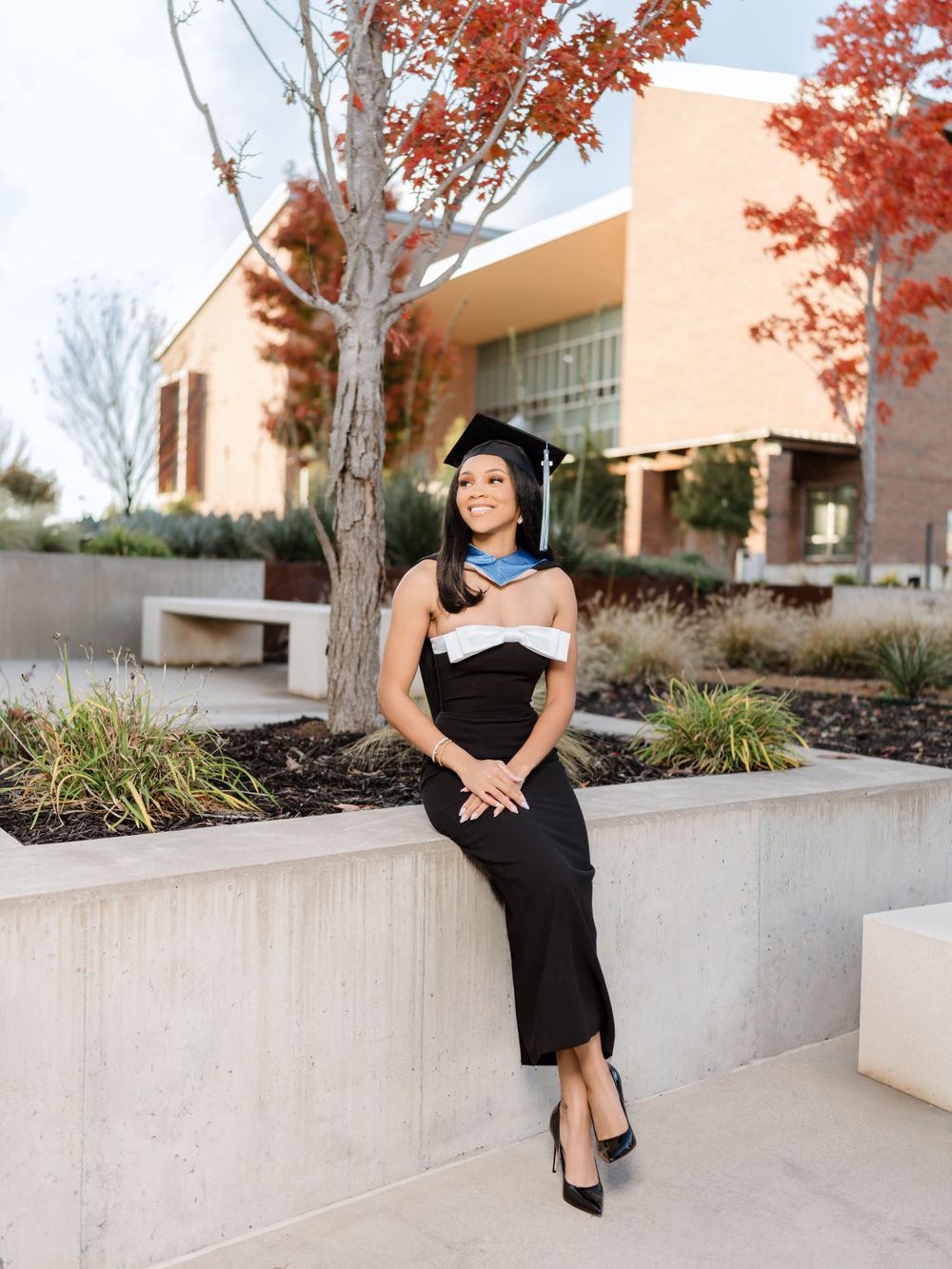 Graduate in black dress and blue stole sits on concrete planter near academic building.