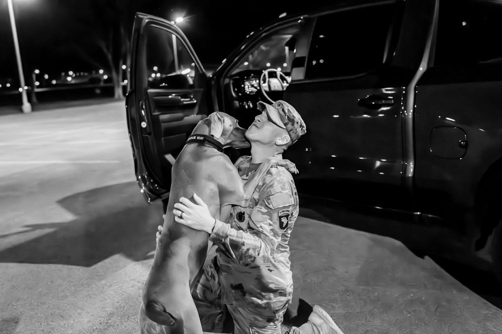 Service member embraces excited dog next to parked vehicle at night.
