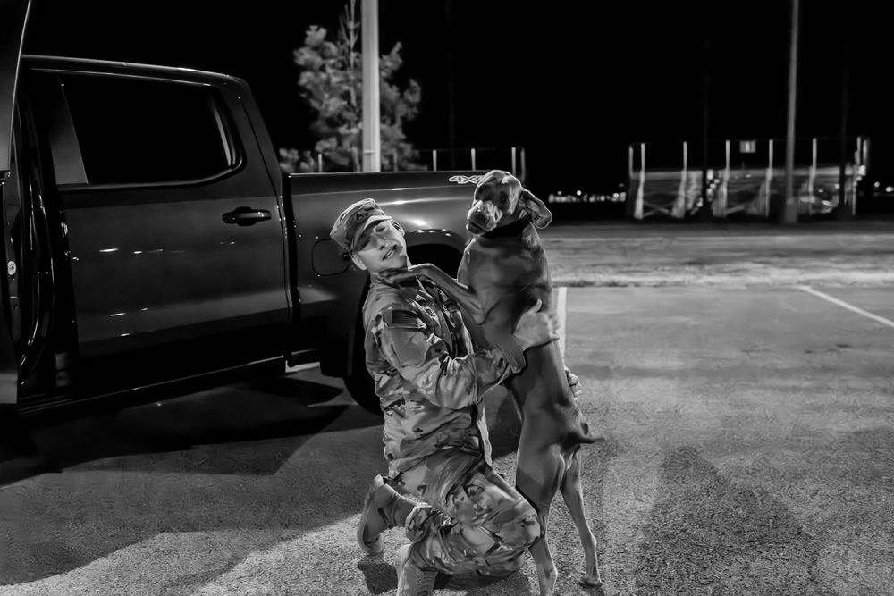 Military service member in uniform joyfully greets excited dog in dark parking lot.
