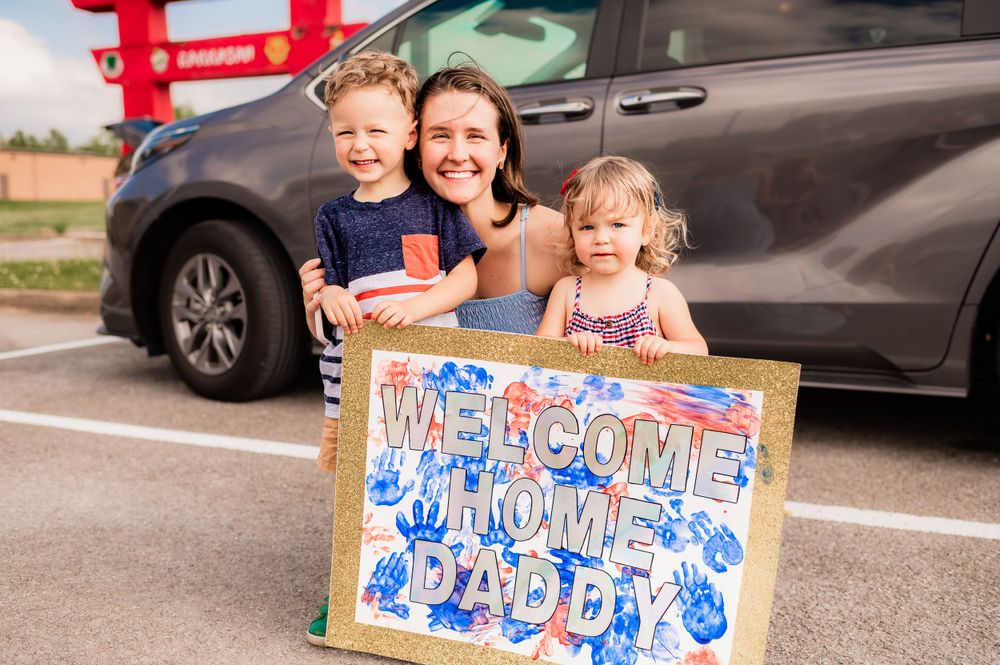 Family poses with handmade welcome home sign in parking lot.
