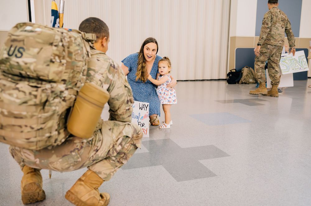 Military homecoming scene with soldier greeting family in indoor venue.