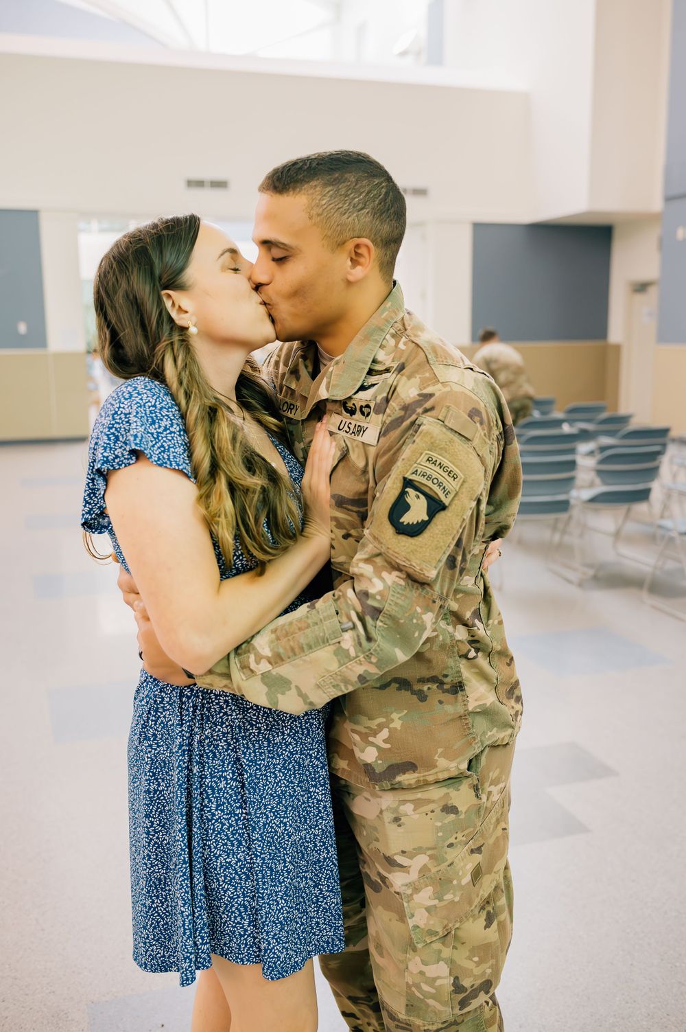 Service member and woman in blue dress share emotional kisses in an empty hallway.