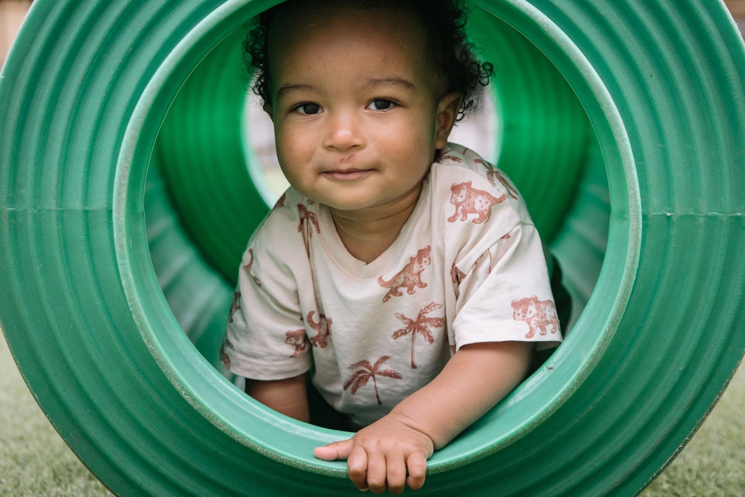 A toddler peeks through a green playground tunnel with a gentle expression.