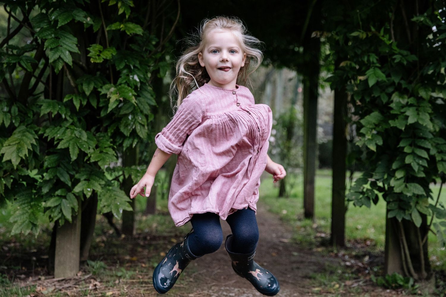 A small child in a pink dress leaps joyfully in a garden setting with trees in the background.