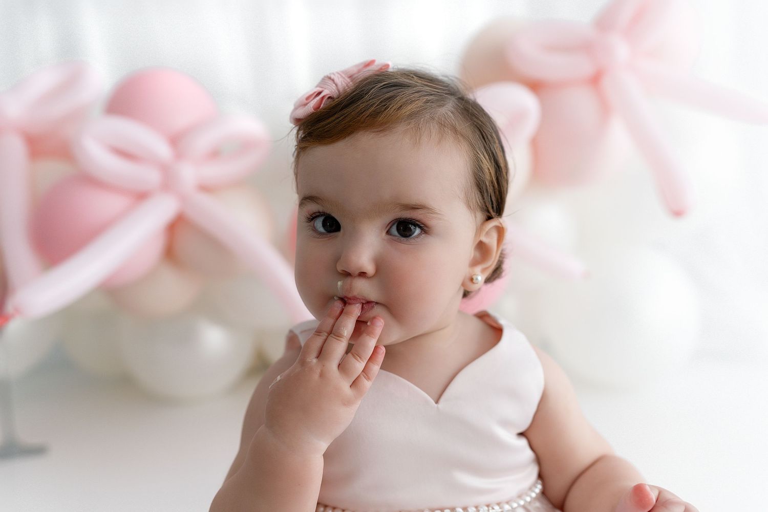 Sweet baby in white dress poses against pink bow background during professional photoshoot.